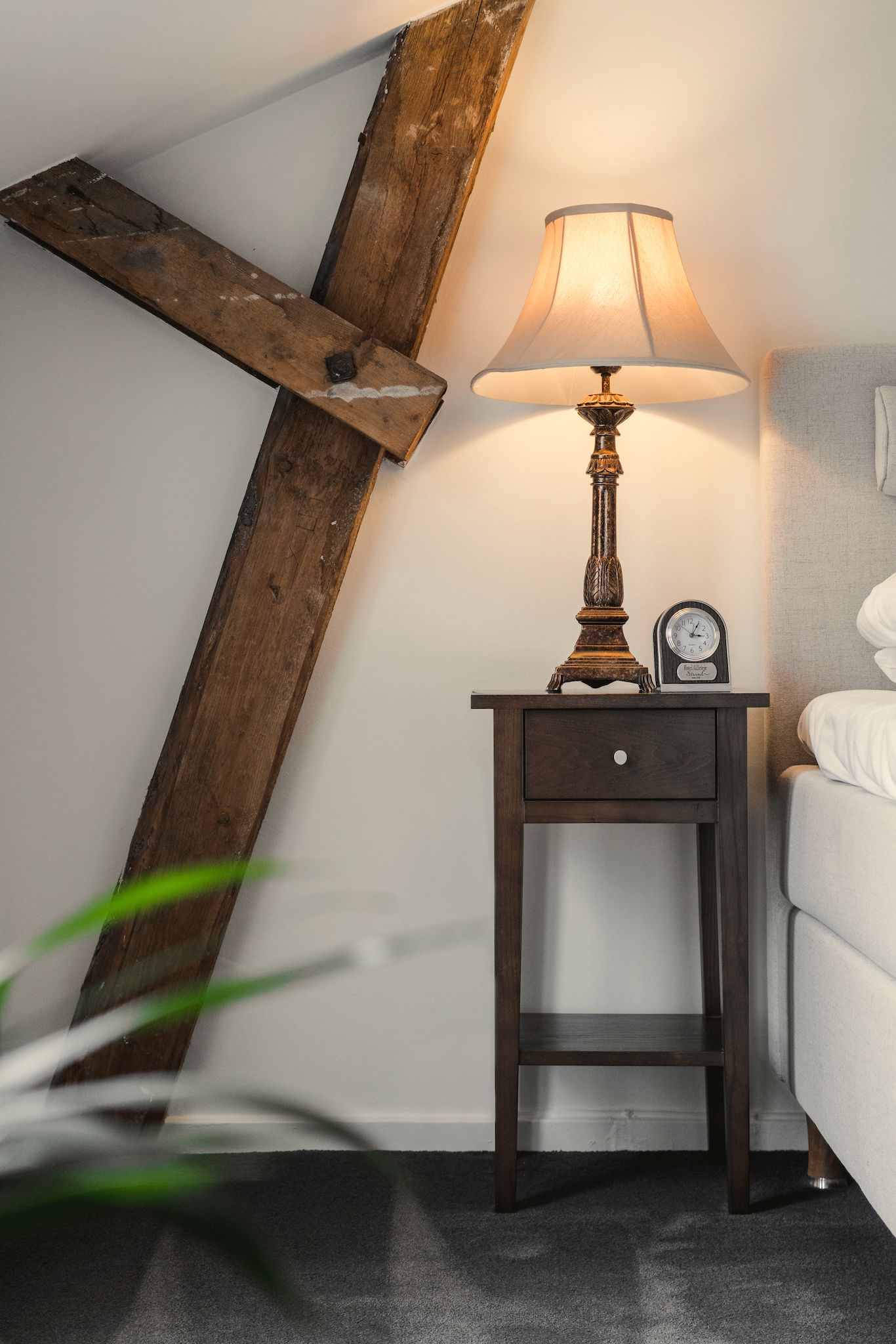 Una mesita de noche de madera con una lámpara y un reloj, ubicada junto a una cama en una habitación con techo inclinado.