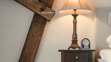 Una mesita de noche de madera con una lámpara y un reloj, ubicada junto a una cama en una habitación con techo inclinado.