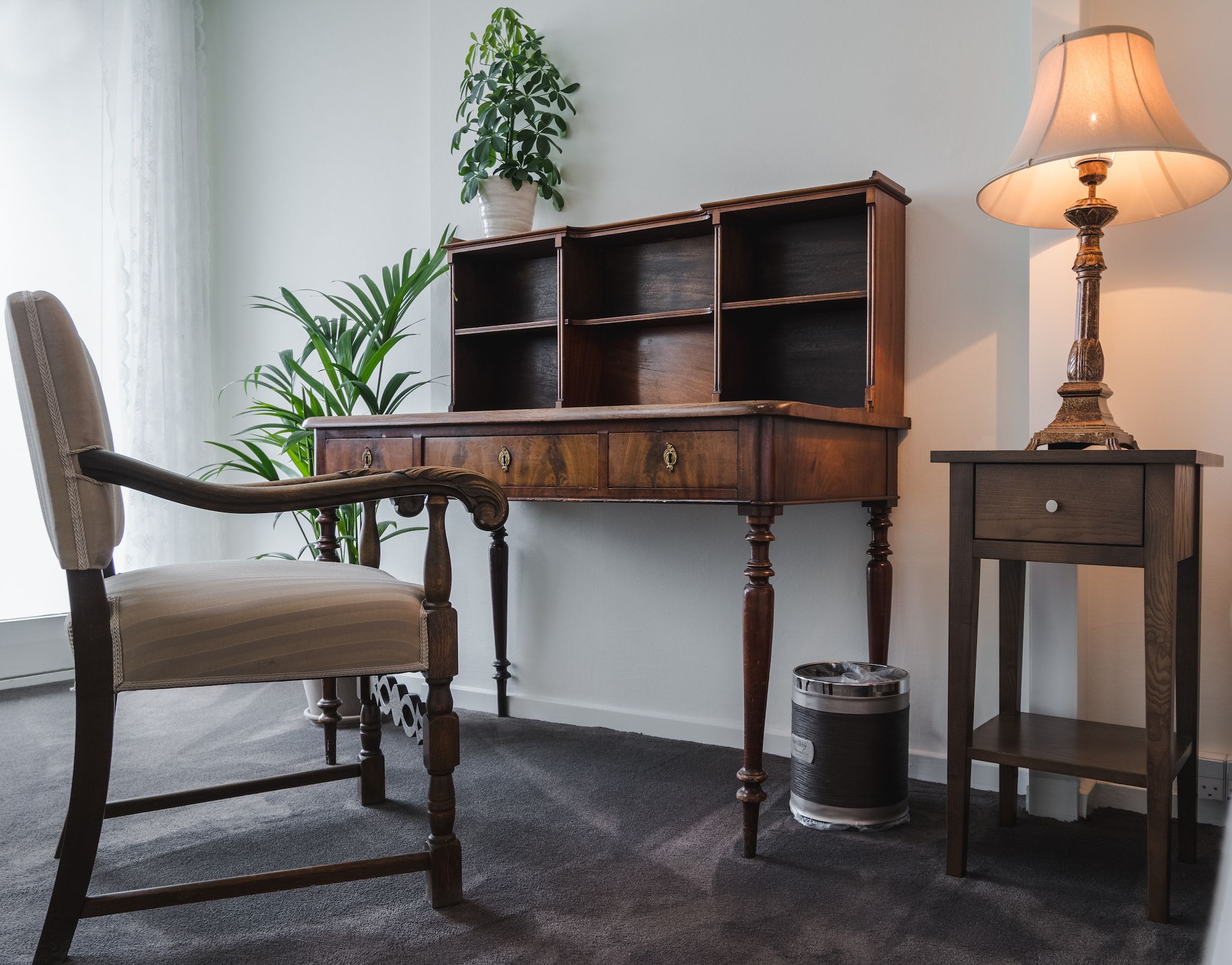 Una habitación con una silla de madera, un escritorio con estantes abiertos, una lámpara y una planta en maceta.