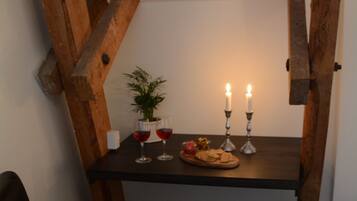 Ein kleiner Holztisch mit Kerzen, Gläsern und einem Teller Essen in einer Ecke eines Raumes mit dunklem Boden und einem schwarzen Ledersessel.