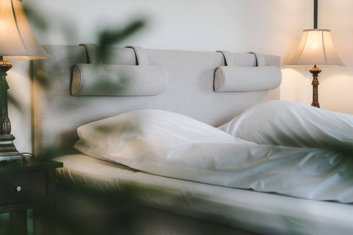 Ein ordentlich bezogenes Bett mit weißen Leinentüchern, ein Kopfteil mit zwei Kissen und je eine Nachttischlampe auf beiden Seiten.