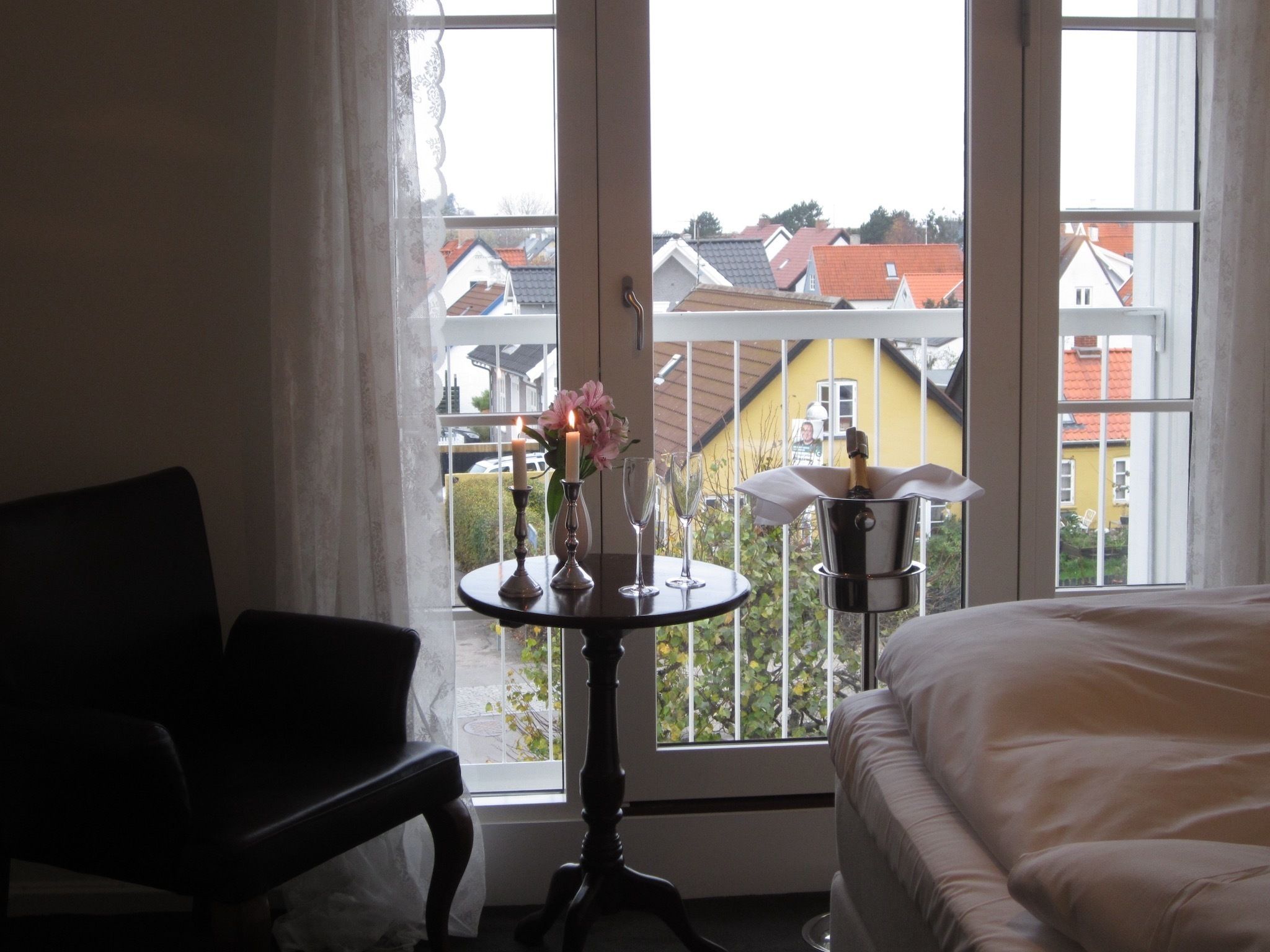 A hotel room with a bed, a chair, a small table with candles and a vase of flowers, and a view of houses through large windows.