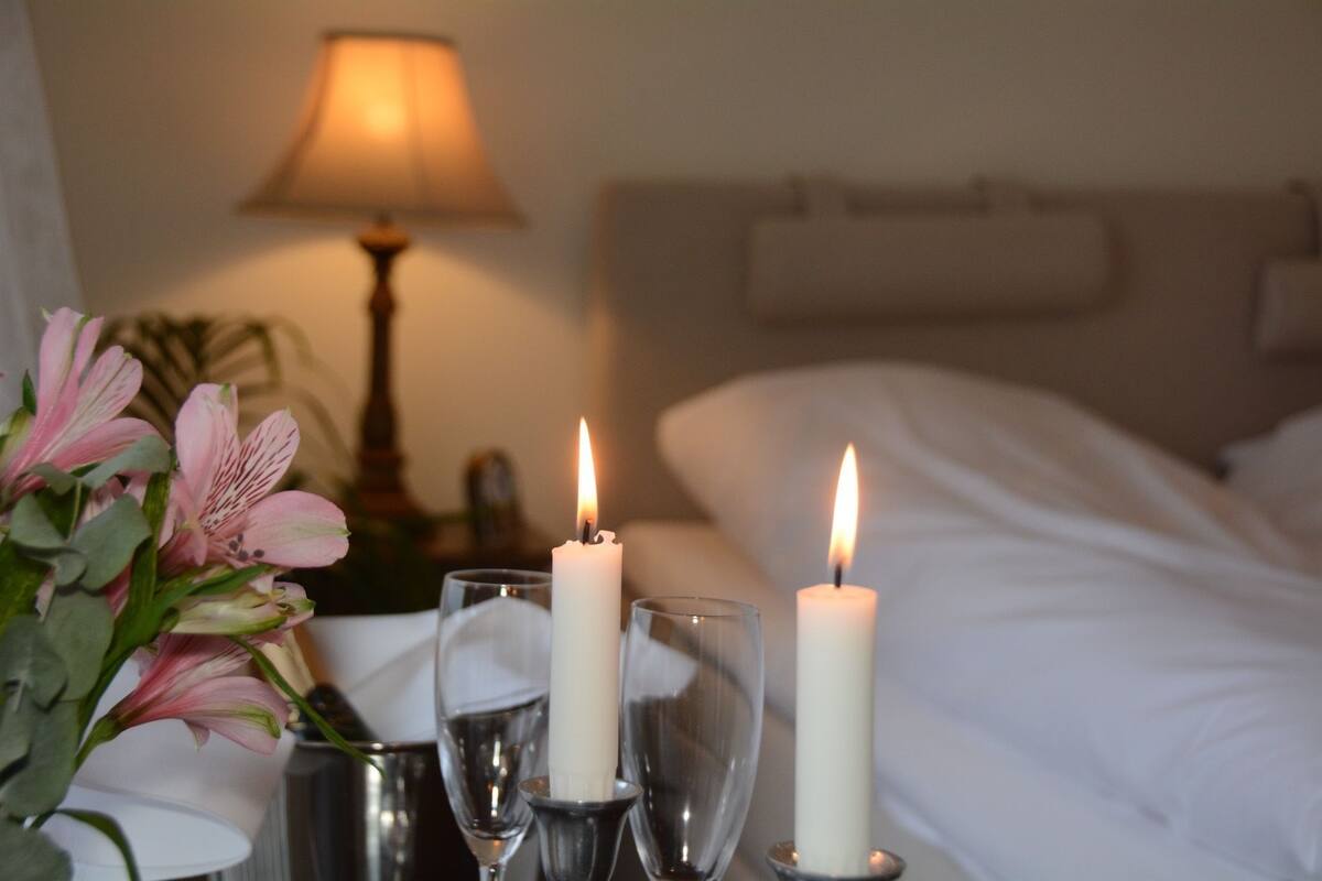 Ein ordentlich bezogenes Bett mit weißen Leinen, zwei brennende Kerzen in klaren Glasgläsern und ein Strauß rosa Blumen auf dem Nachttisch.