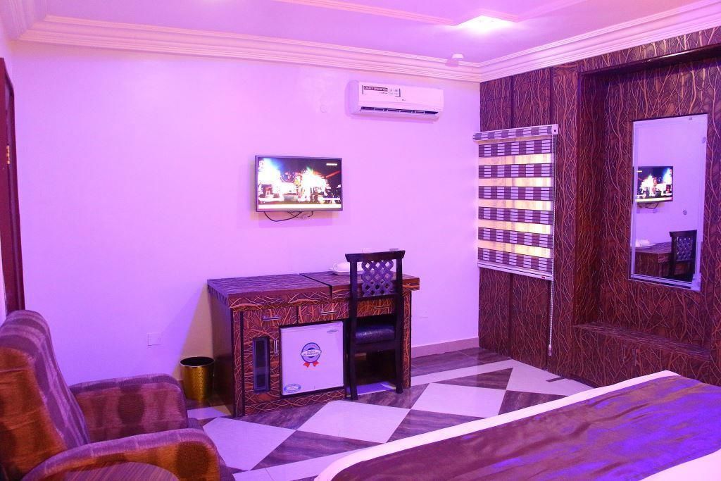 A hotel room with a checkered floor, a bed, a desk with a chair, a TV mounted on the wall, and an air conditioning unit.