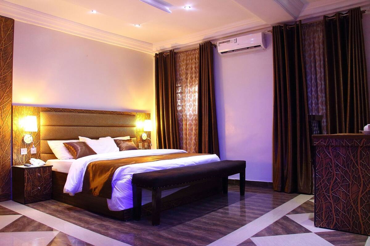 A hotel room with a large bed, bedside lamps, an air conditioning unit, a bench, and a patterned wall.