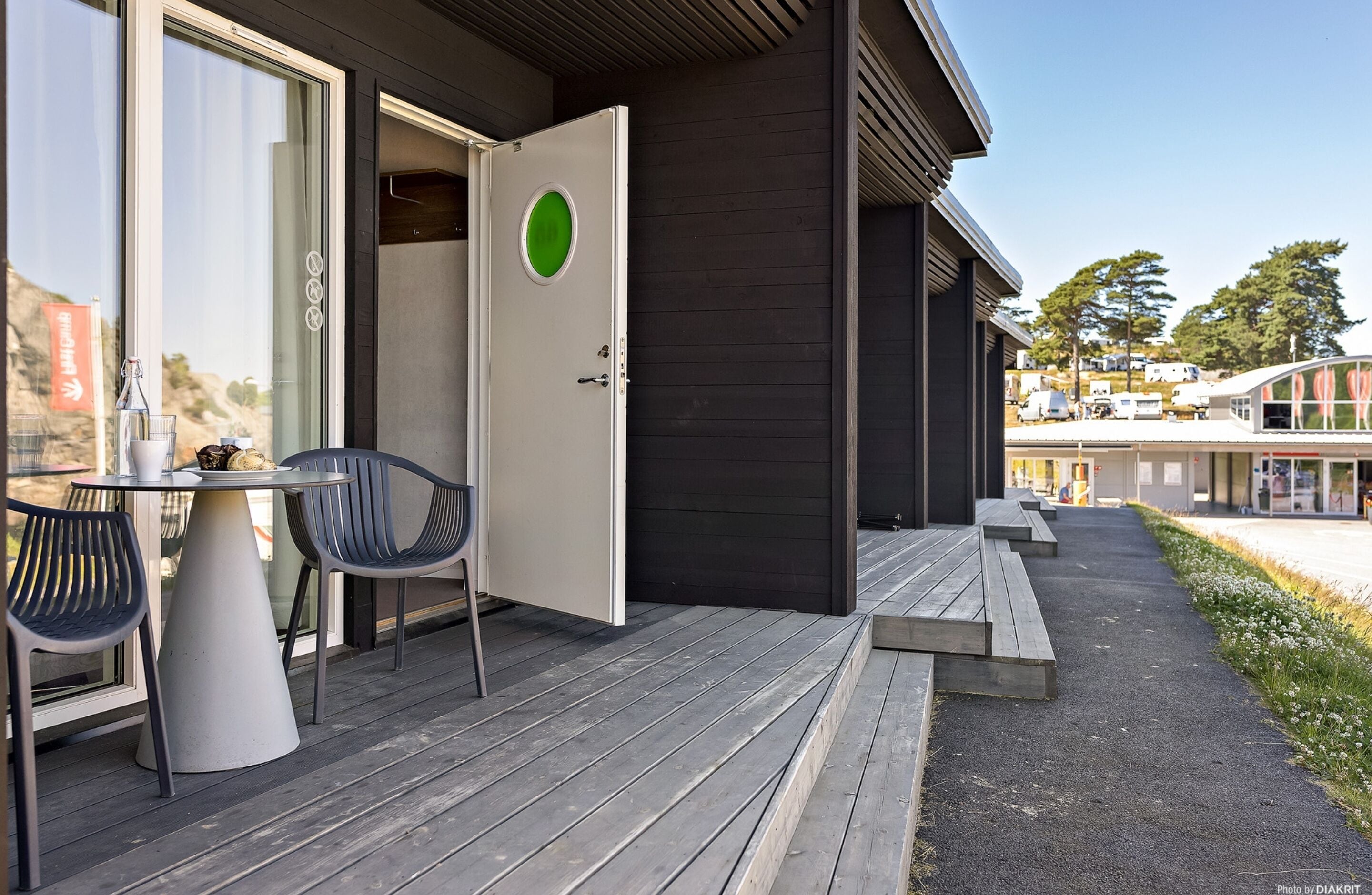 A wooden deck with a table and chairs, a door with a green circle, and a view of a building with a glass facade.