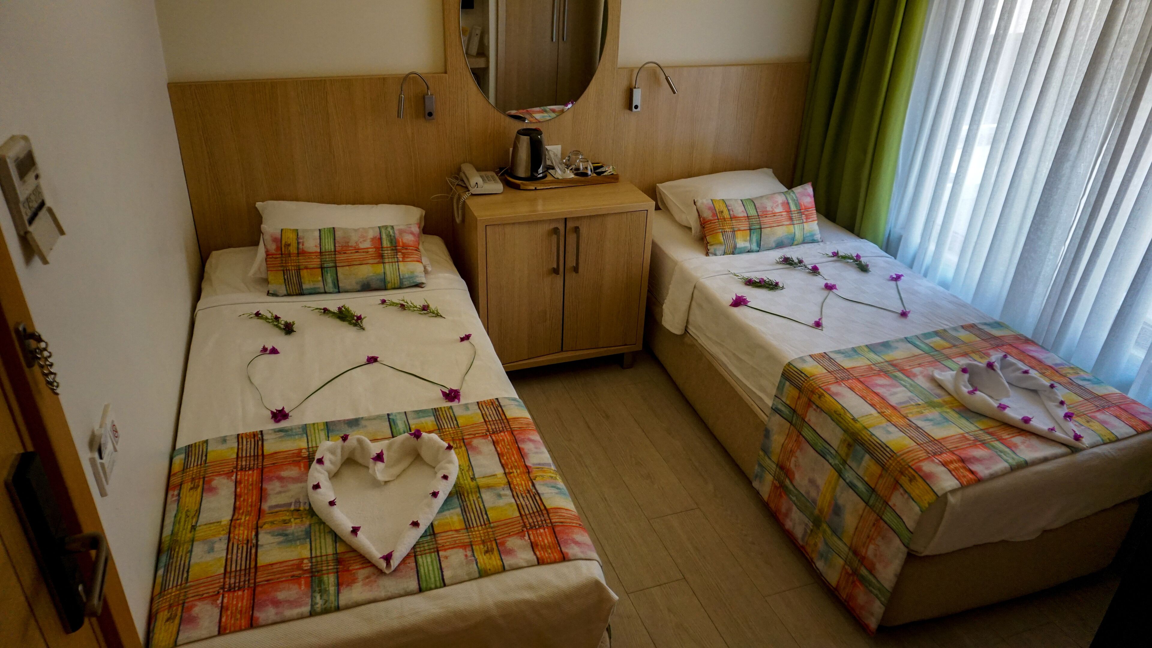 A hotel room with two beds, each with a colorful bedspread and heart-shaped decorations. There is a wooden wardrobe, a bedside table with a kettle, and a mirror above the bed.