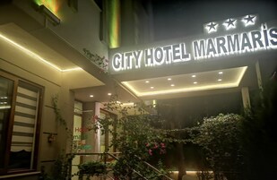 The exterior of City Hotel Marmaris with a prominent illuminated sign, a staircase, and lush greenery.