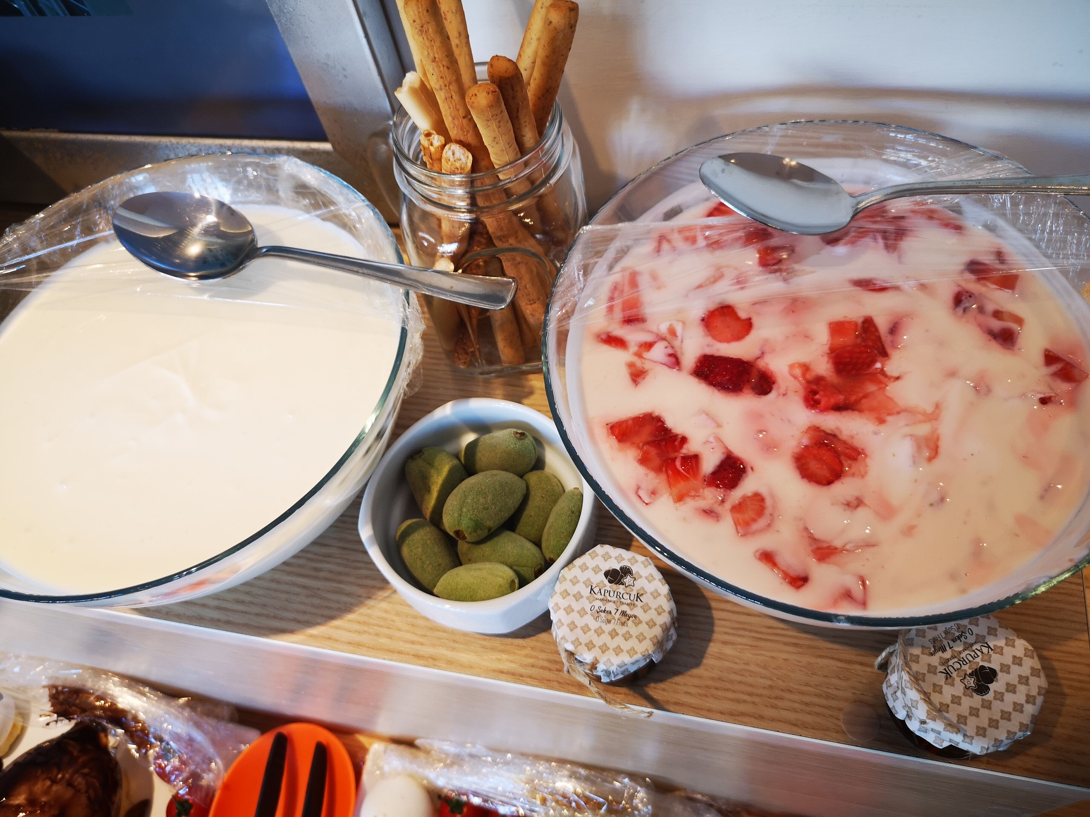 Un plateau avec deux bols de nourriture : l’un contient une substance blanche et crémeuse, et l’autre, un mélange au goût de fraise. On y trouve également quelques noix vertes et un bocal qui semble contenir des biscuits ou des gâteaux secs.