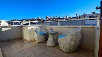 Una terraza en la azotea con dos sillas de mimbre y una pequeña mesa, con vistas a un paisaje urbano que incluye edificios y montañas.