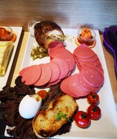 A plate of sliced meat, a boiled egg, grilled bread, and tomatoes on a wooden table.