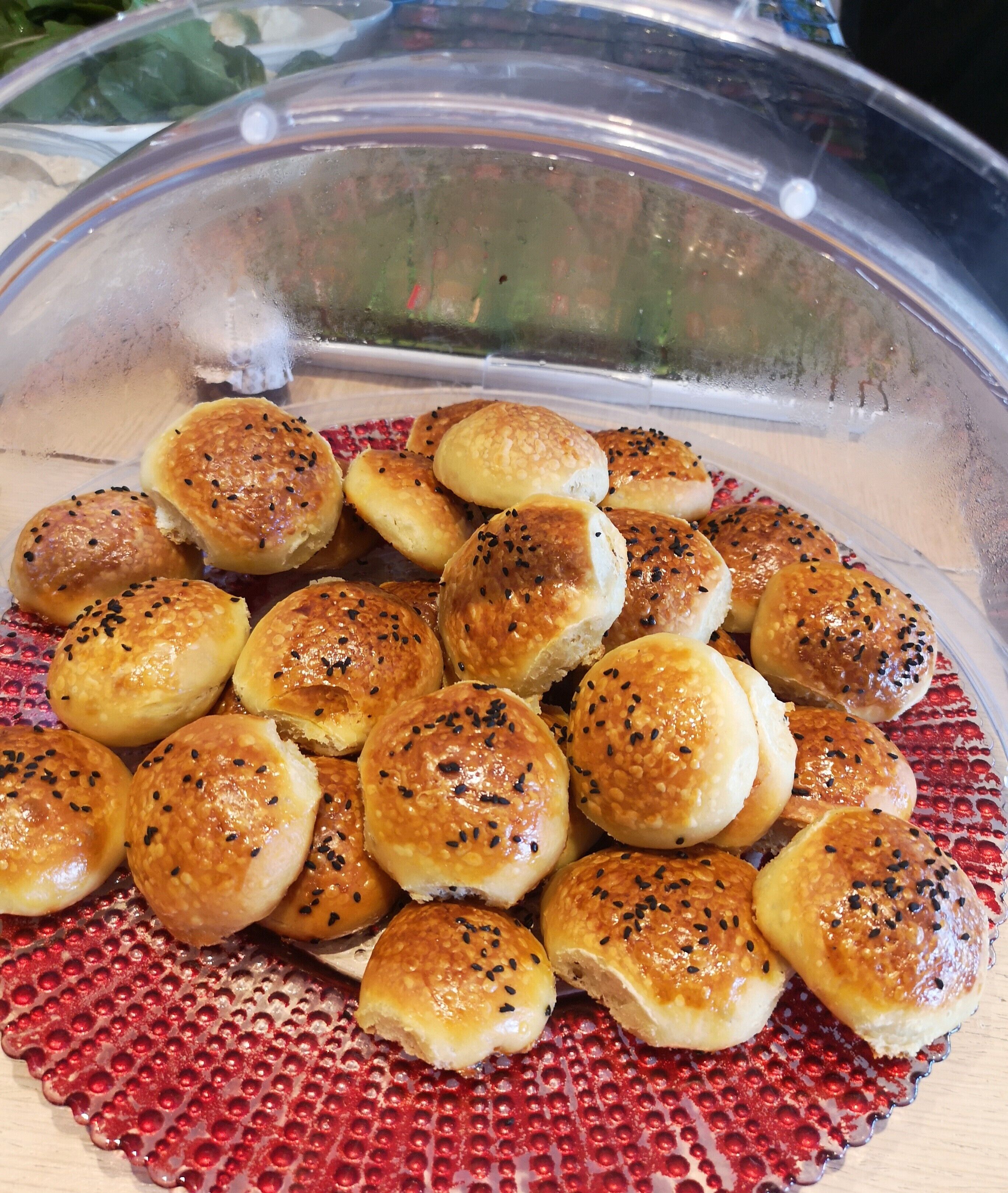 A clear plastic container filled with round, golden-brown bread rolls topped with black sesame seeds, placed on a red textured plate.