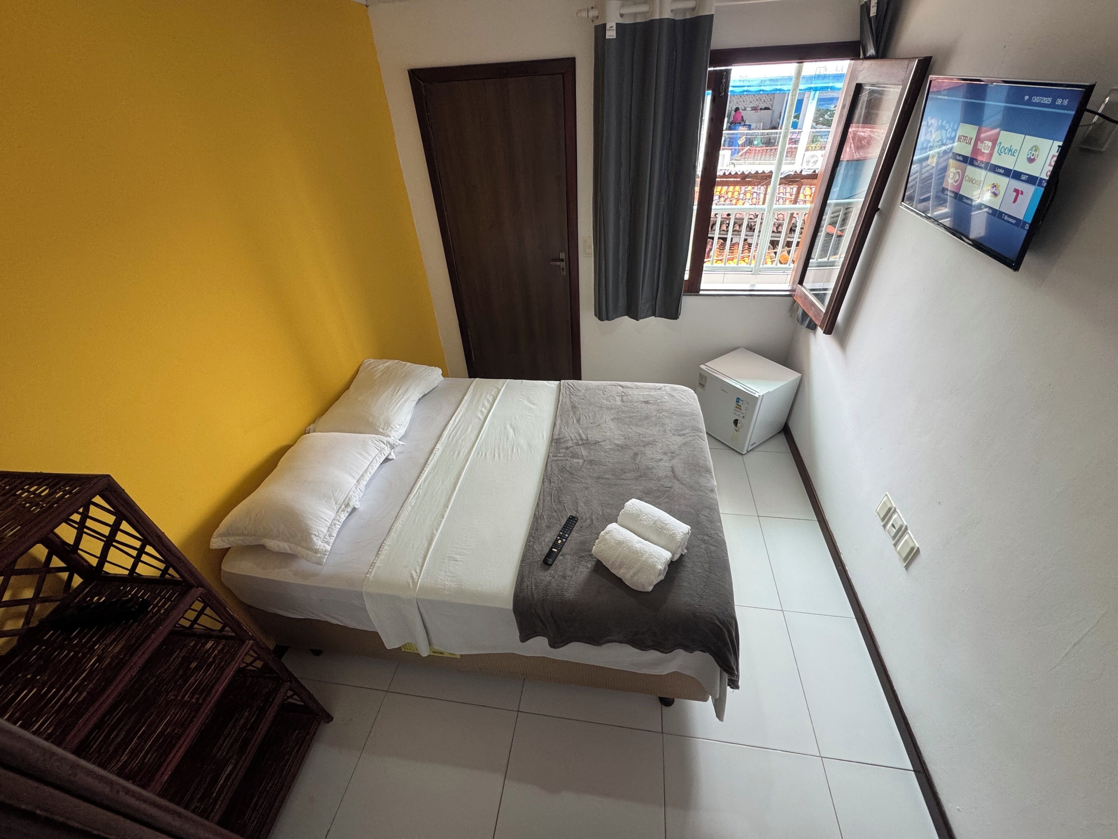A bedroom with a bed, a small refrigerator, a window with curtains, and a wall-mounted TV.