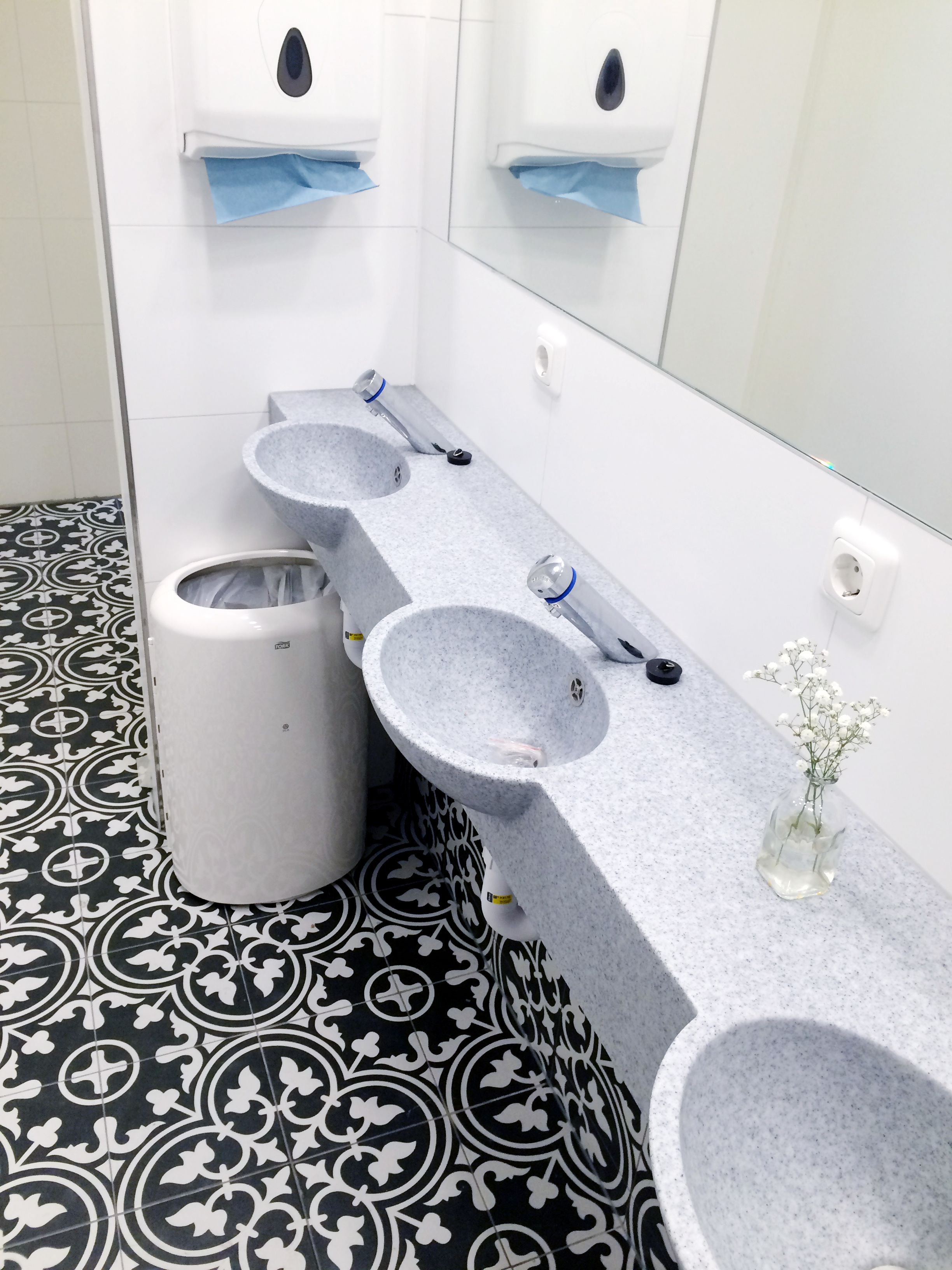 A bathroom with three sinks, a trash bin, and a vase with flowers.