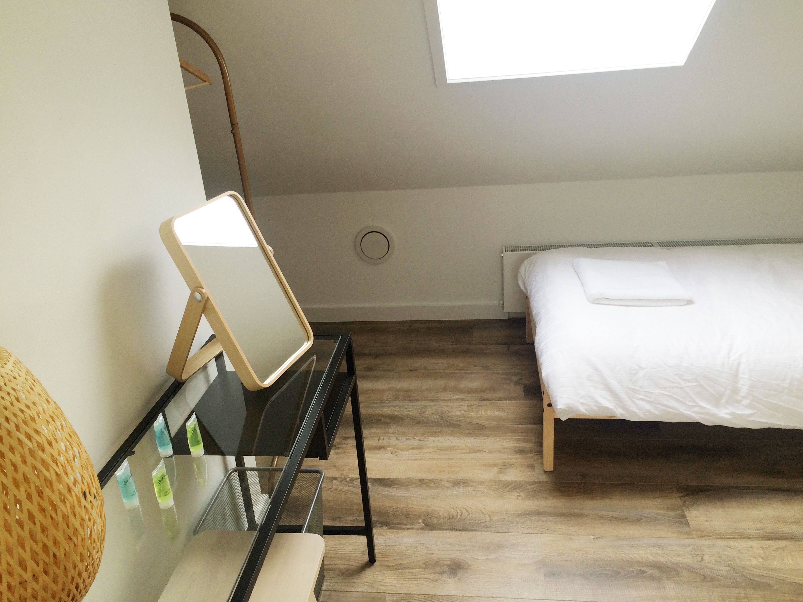 A bedroom with a bed, a desk with a mirror, a chair, and a skylight.