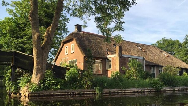 A house with a thatched roof, a brick chimney, and a reflection in the water.