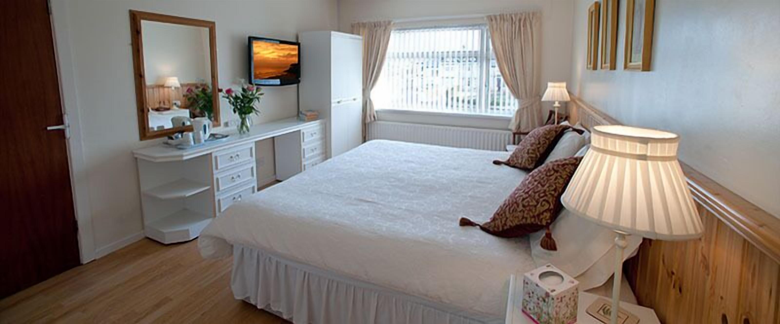 A neatly arranged bedroom with a bed, bedside lamps, a dresser, a television, and a window with curtains.