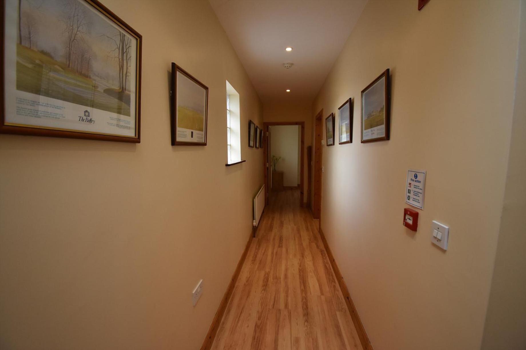 A hallway with framed pictures on the walls, a wooden floor, and a fire action sign.