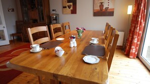 Una sala da pranzo con un tavolo in legno apparecchiato per quattro persone, circondato da sedie. Si notano due quadri alle pareti, un armadio con vetrine e una finestra con tende.