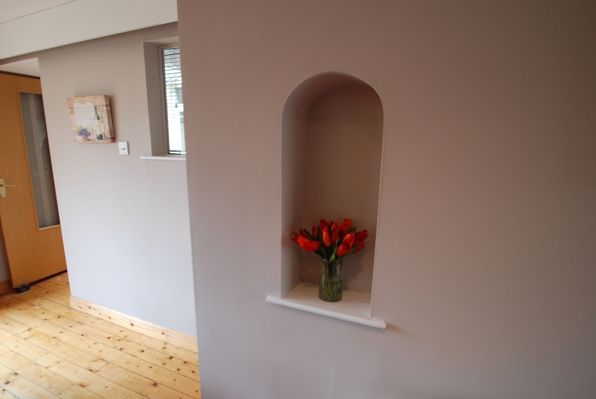 Un pasillo con suelo de madera, una puerta, un ventanal pequeño y un nicho en la pared con un jarrón de flores rojas.