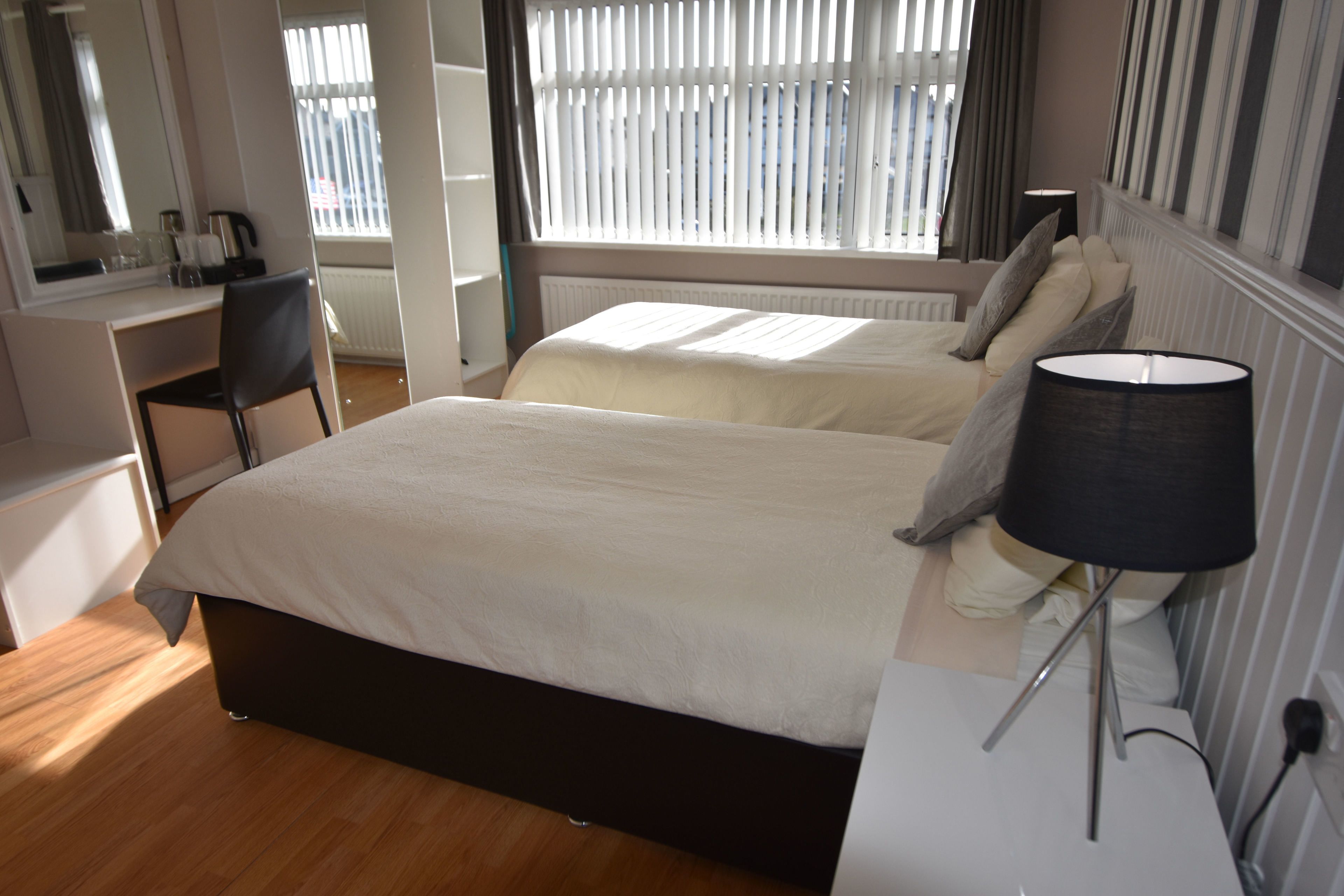 A hotel room with two beds, a desk, a chair, a lamp, and a window with blinds.
