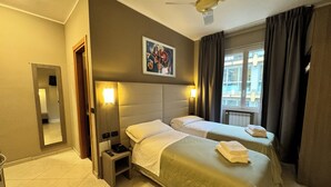 Een hotelkamer met twee bedden, een plafondventilator, een schilderij, een raam met gordijnen en uitzicht op gebouwen.