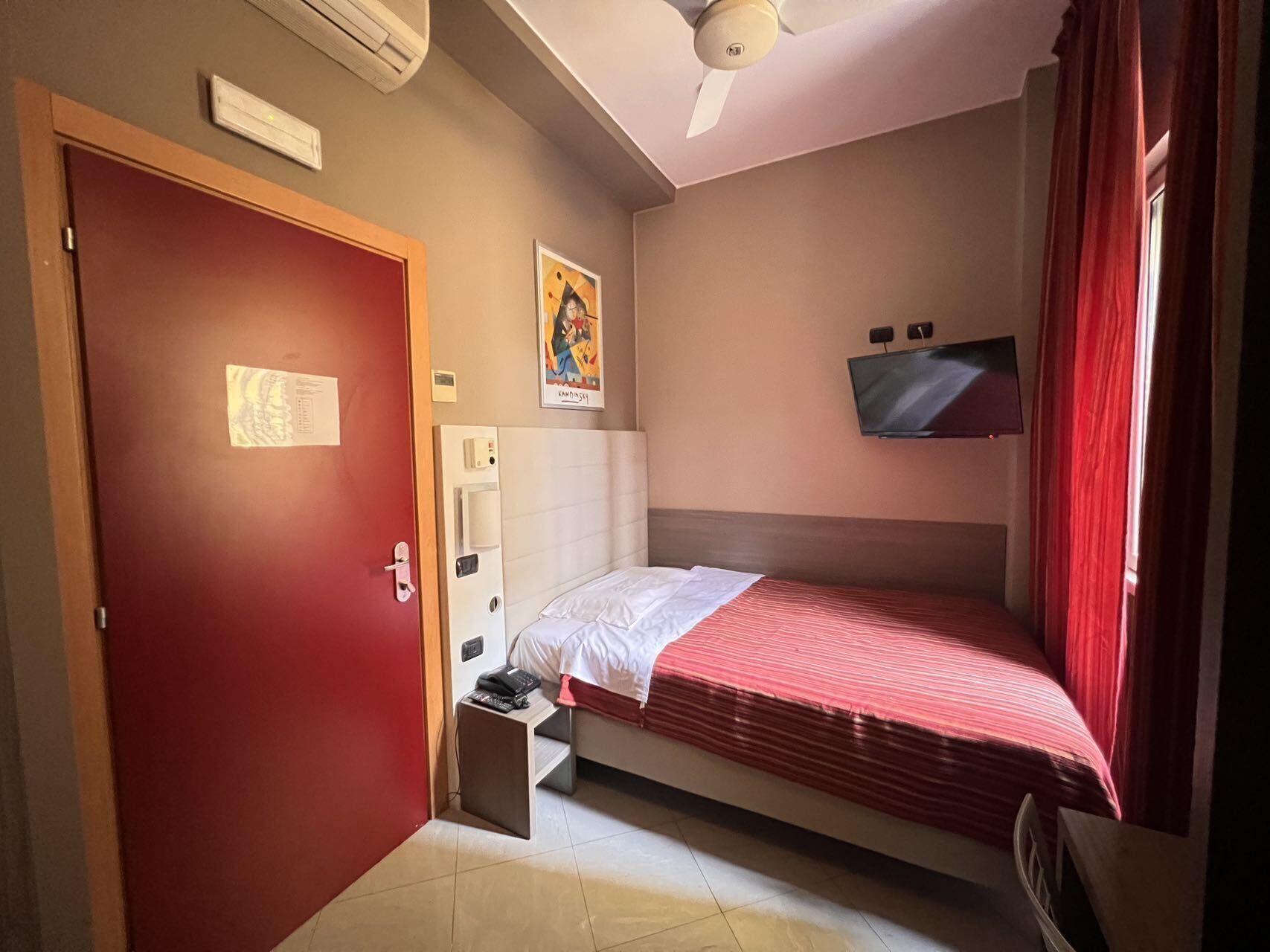 A small, well-lit room with a single bed, a TV mounted on the wall, a door with a handle, and a ceiling fan.