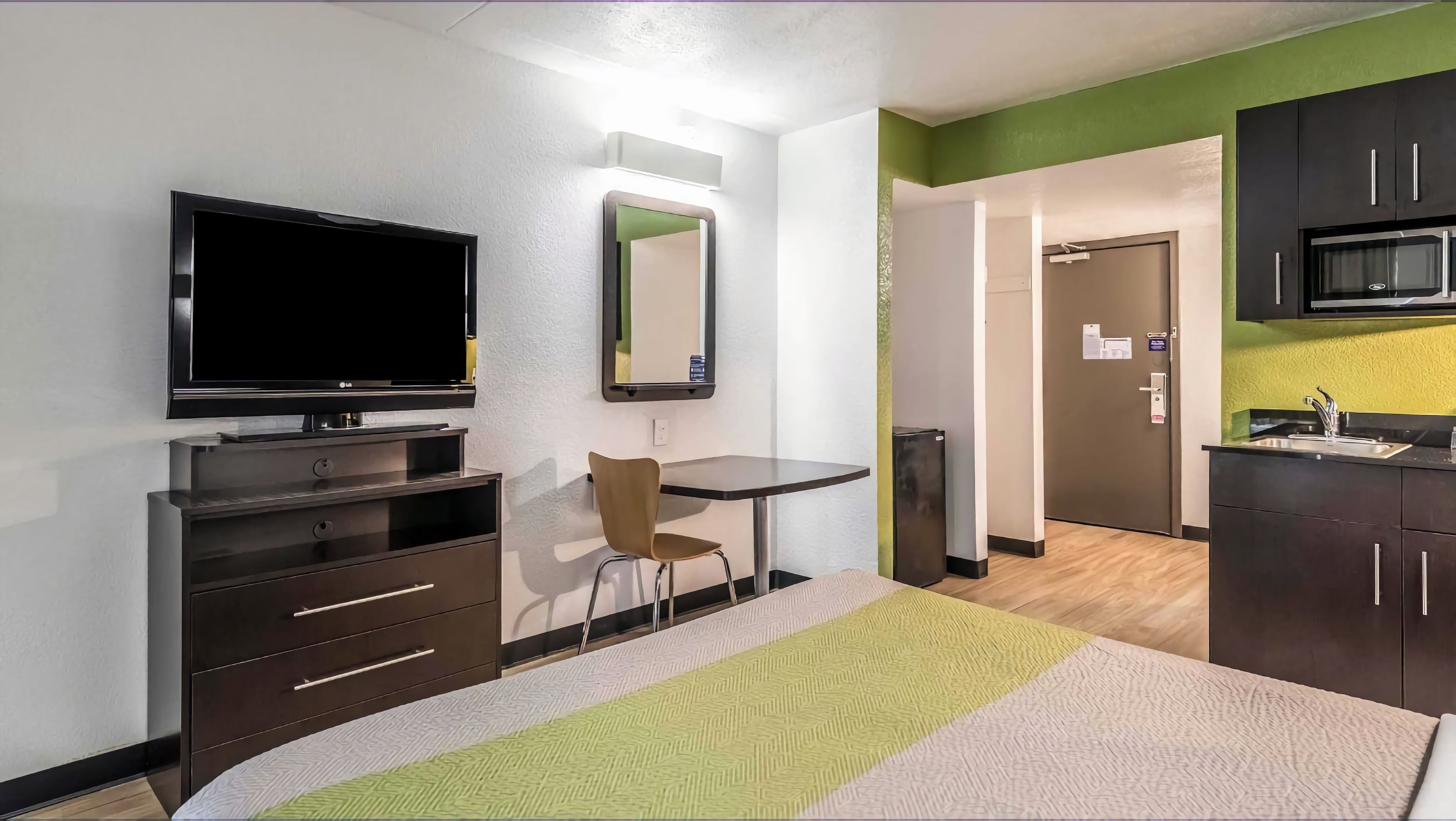 A hotel room with a bed, a desk, a chair, a television, a mirror, a microwave, and a kitchenette.