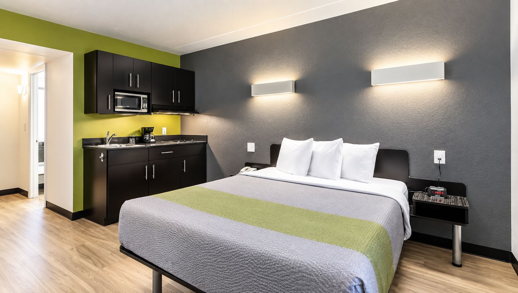 A hotel room with a bed, a bedside table with a clock, a wall-mounted light, and a kitchenette with a microwave.