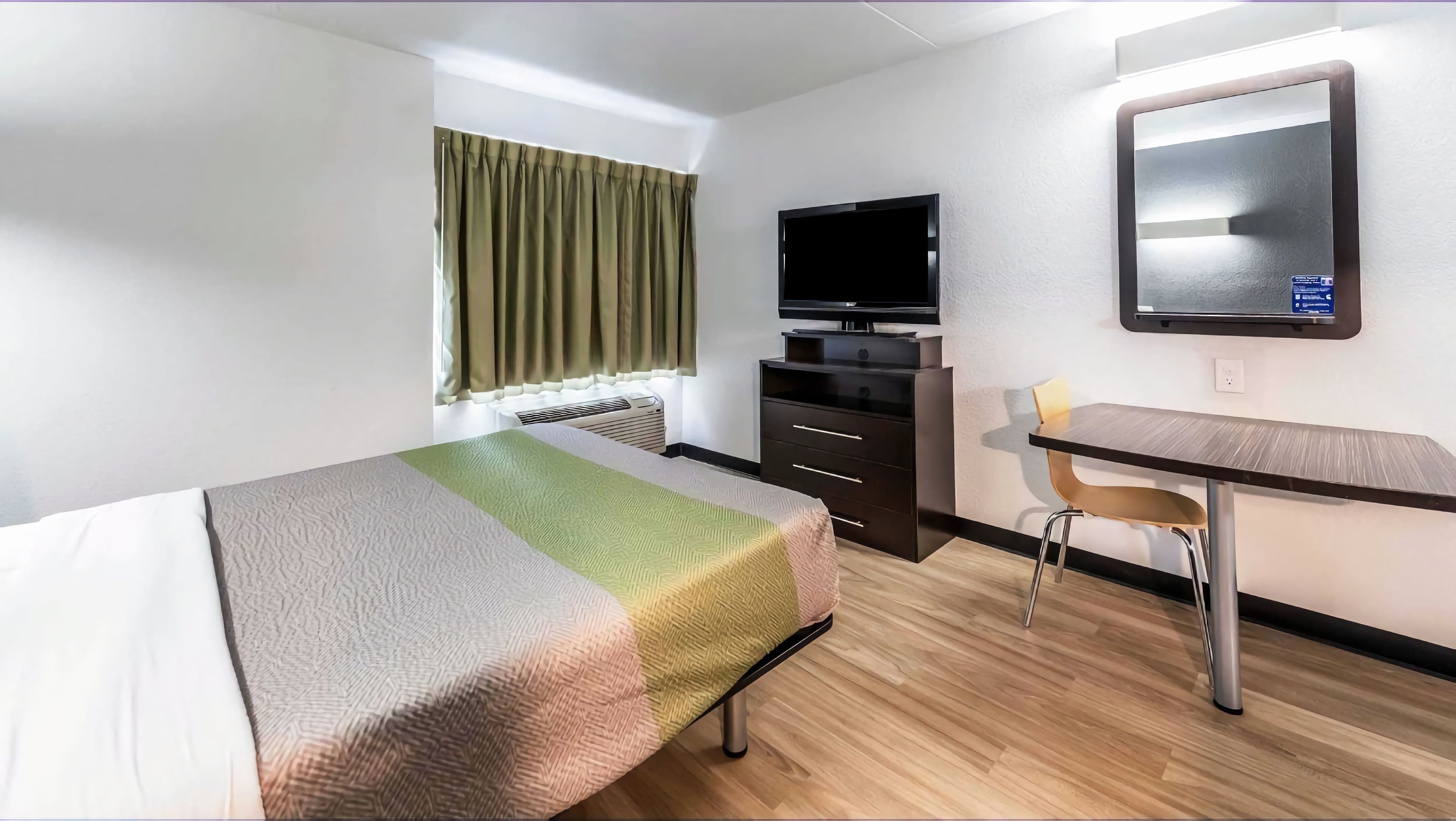 A hotel room with a bed, a desk with a chair, a television, and a window with curtains.