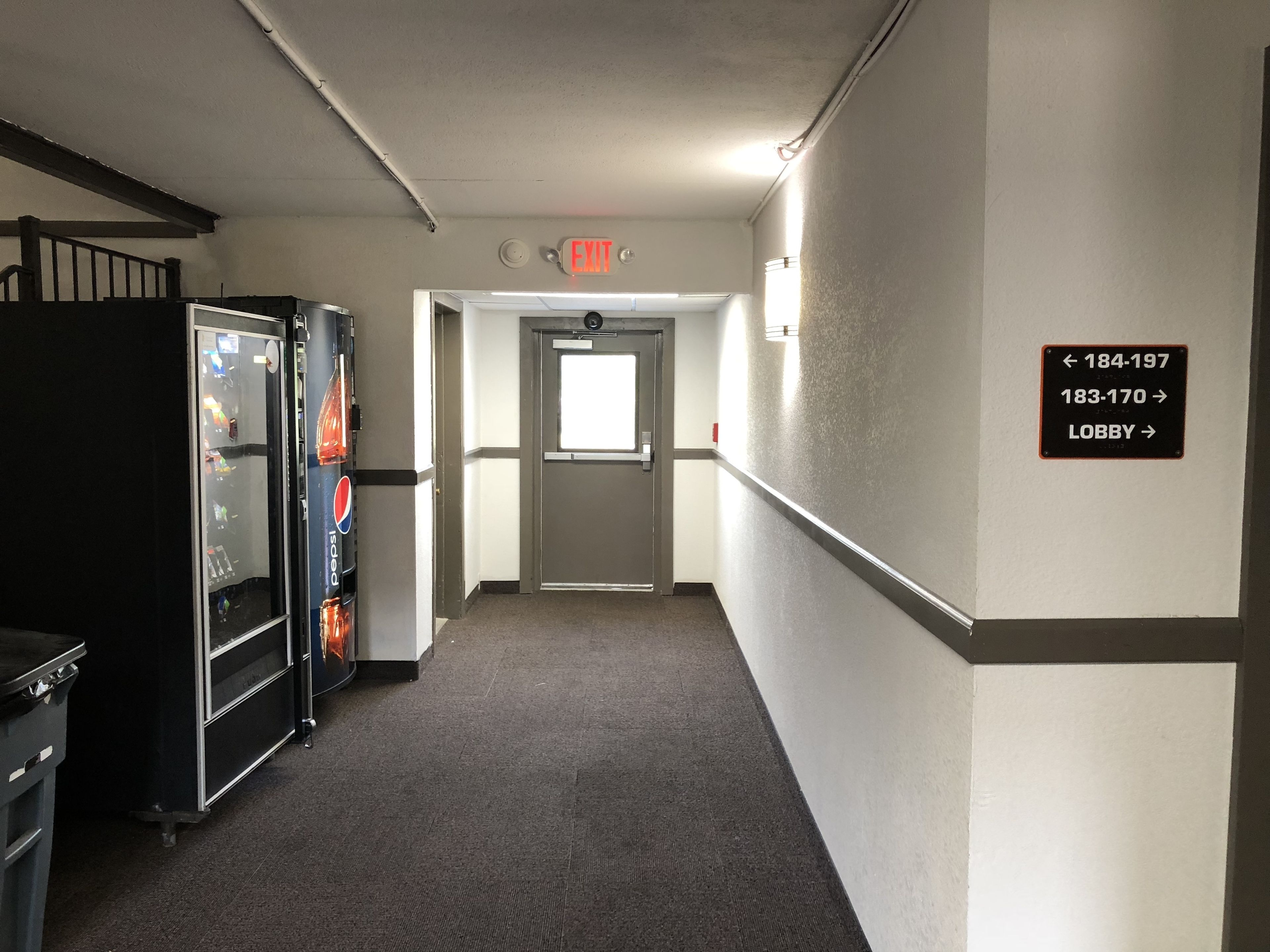 A hallway with a vending machine, an exit sign, and directional signs for rooms and the lobby.