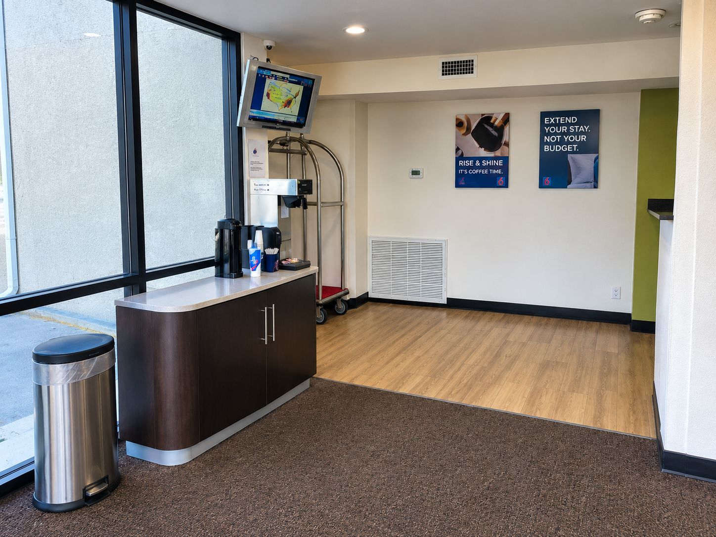 A hotel reception area with a coffee machine, a trash bin, a monitor, and a poster on the wall.