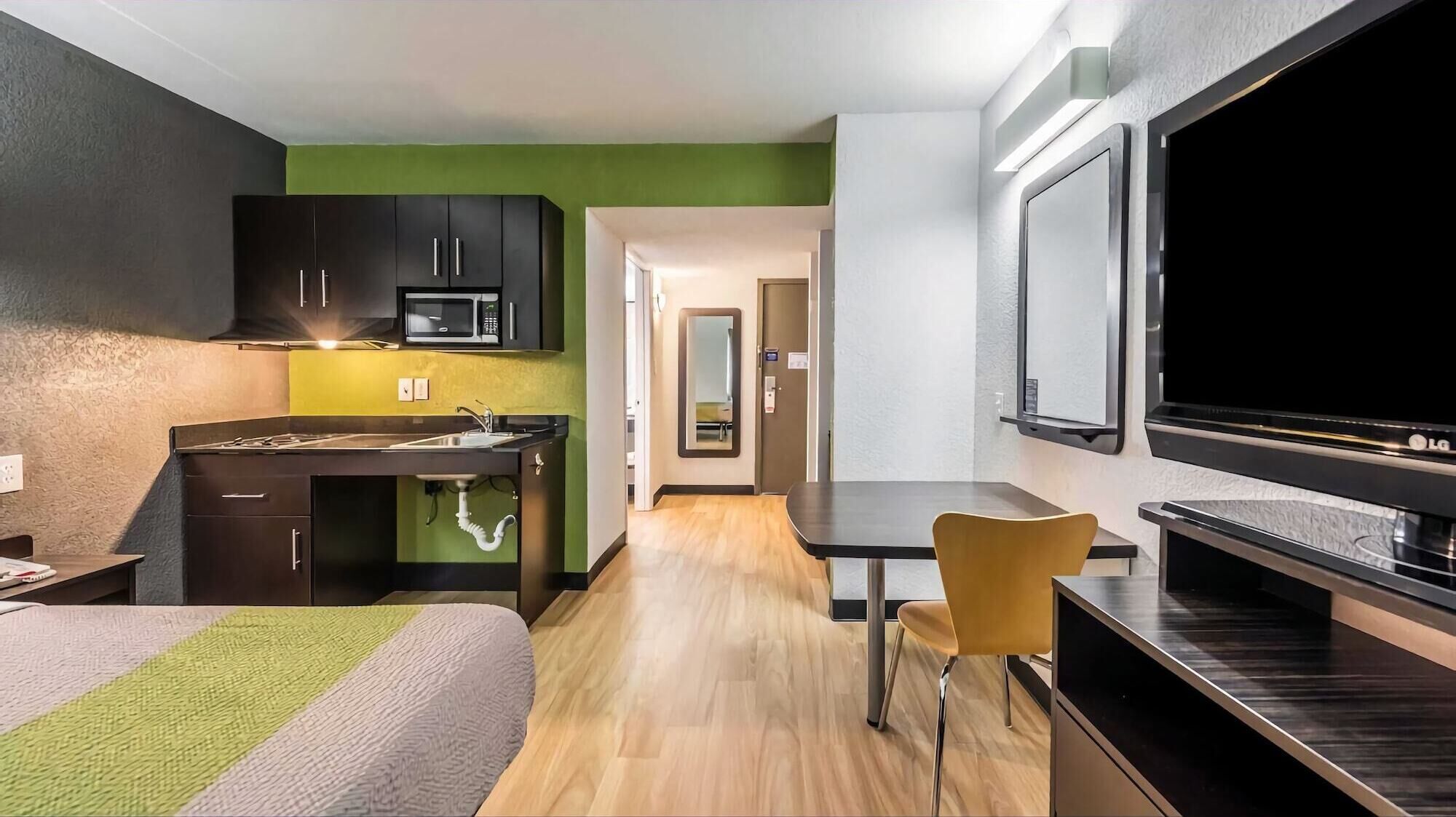 A compact hotel room with a kitchenette, a bed, a TV, and a chair.