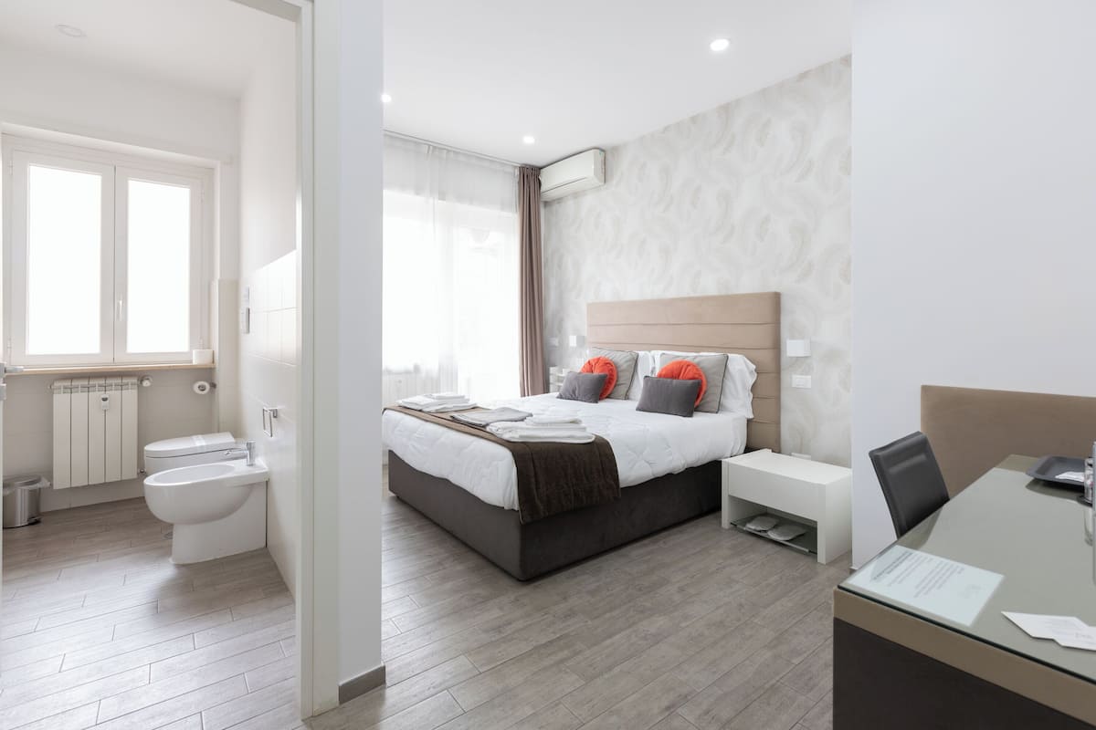 A modern hotel room with a large bed, bedside tables, a desk, and a bathroom with a toilet and sink.
