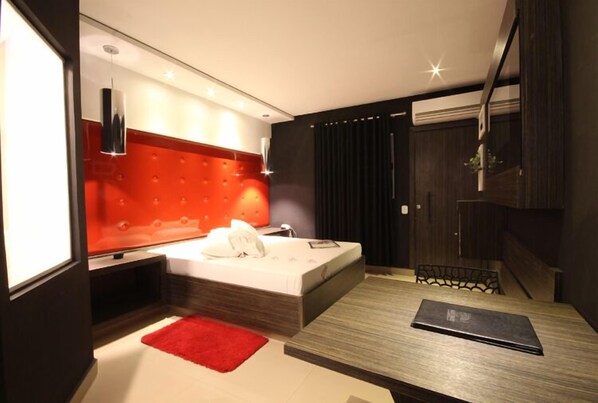 Un dormitorio moderno con cabecera roja, una cama, una mesita de noche, una alfombra roja y un ordenador portátil sobre la mesa.