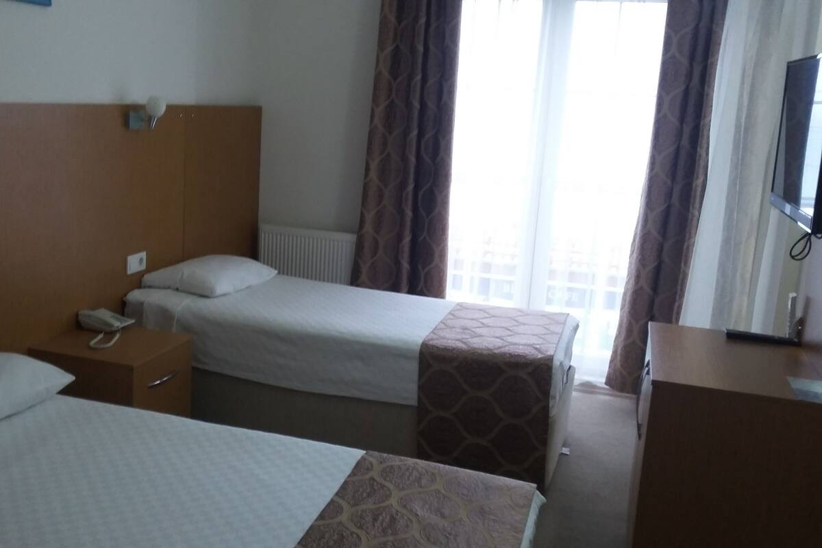 A hotel room with two beds, a bedside table with a phone, a radiator, and a window with curtains.