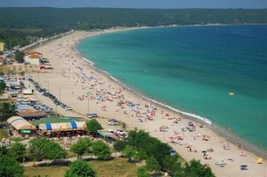 A beach with numerous umbrellas and people, a clear blue sea, and buildings along the shore.