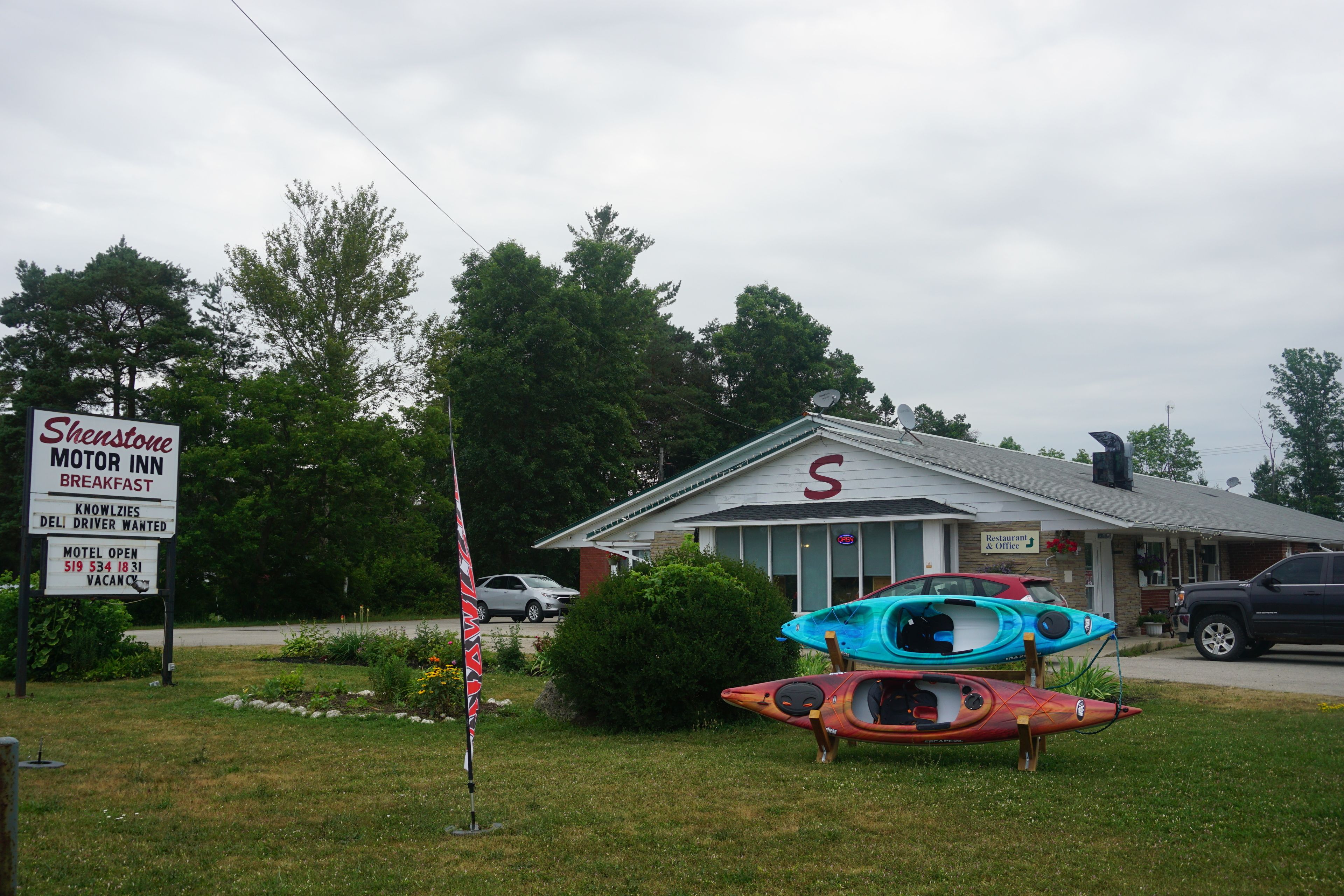 A motel named Shenstone Motor Inn with a signboard indicating breakfast, a restaurant, and a deli. There are kayaks displayed outside.