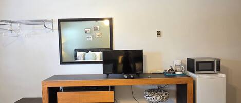 Habitación de hotel compacta con escritorio de madera, televisor de pantalla plana, refrigerador blanco pequeño y lámpara de pie con estampado.