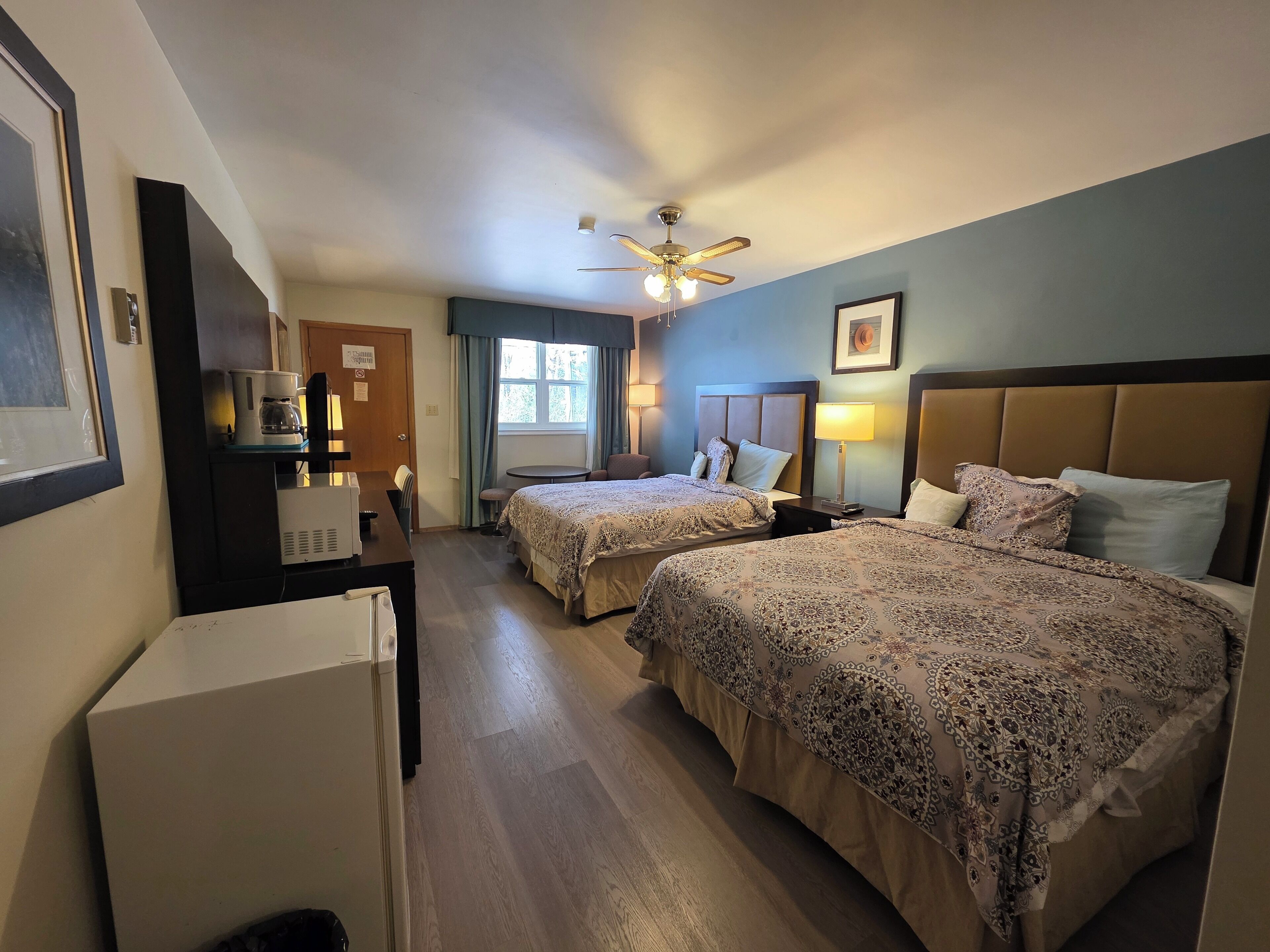 A hotel room with two beds, a desk with a coffee maker, a ceiling fan, and a window with curtains.
