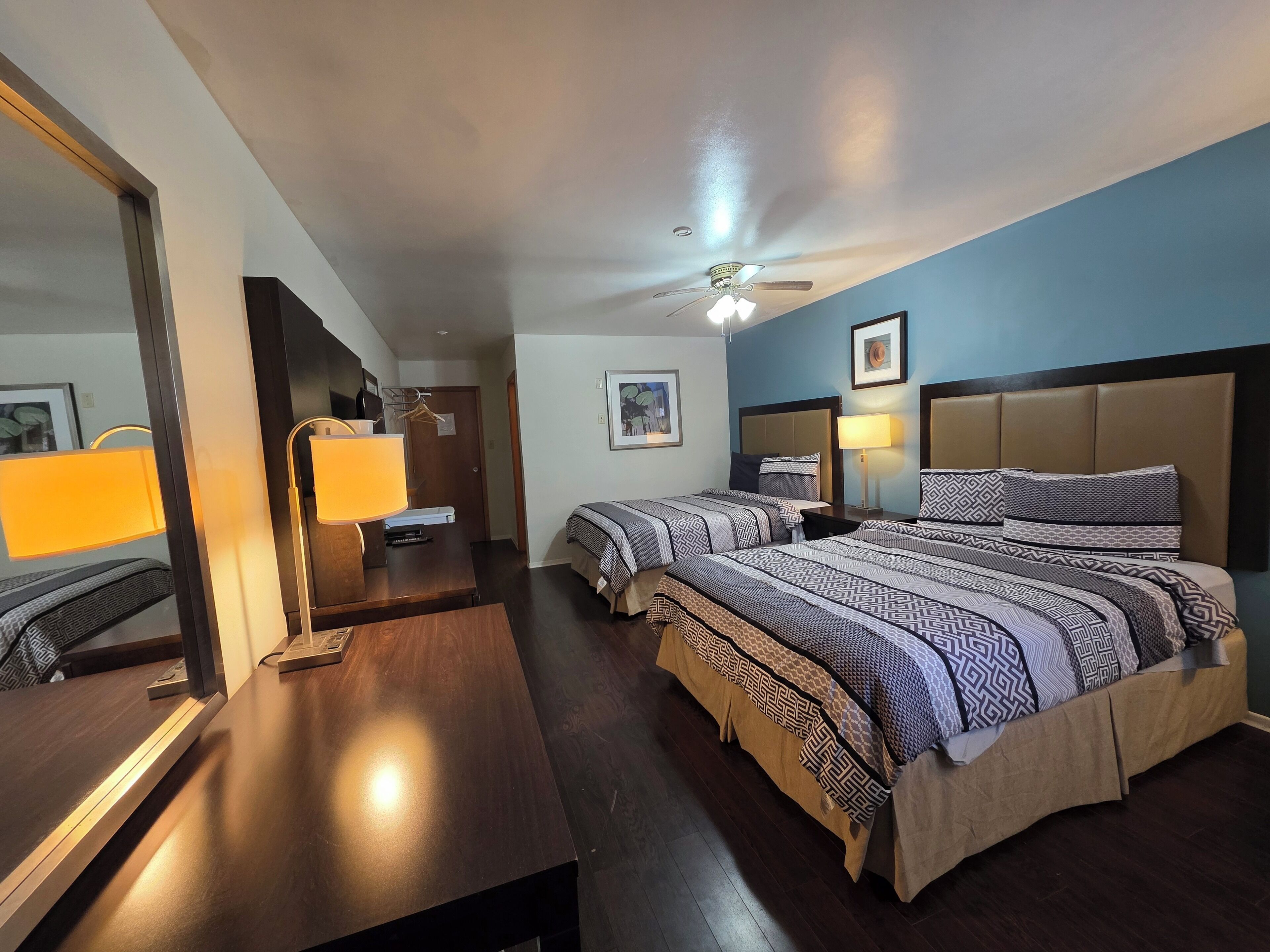 A hotel room with two beds, a desk, a mirror, a ceiling fan, and a wall-mounted lamp.