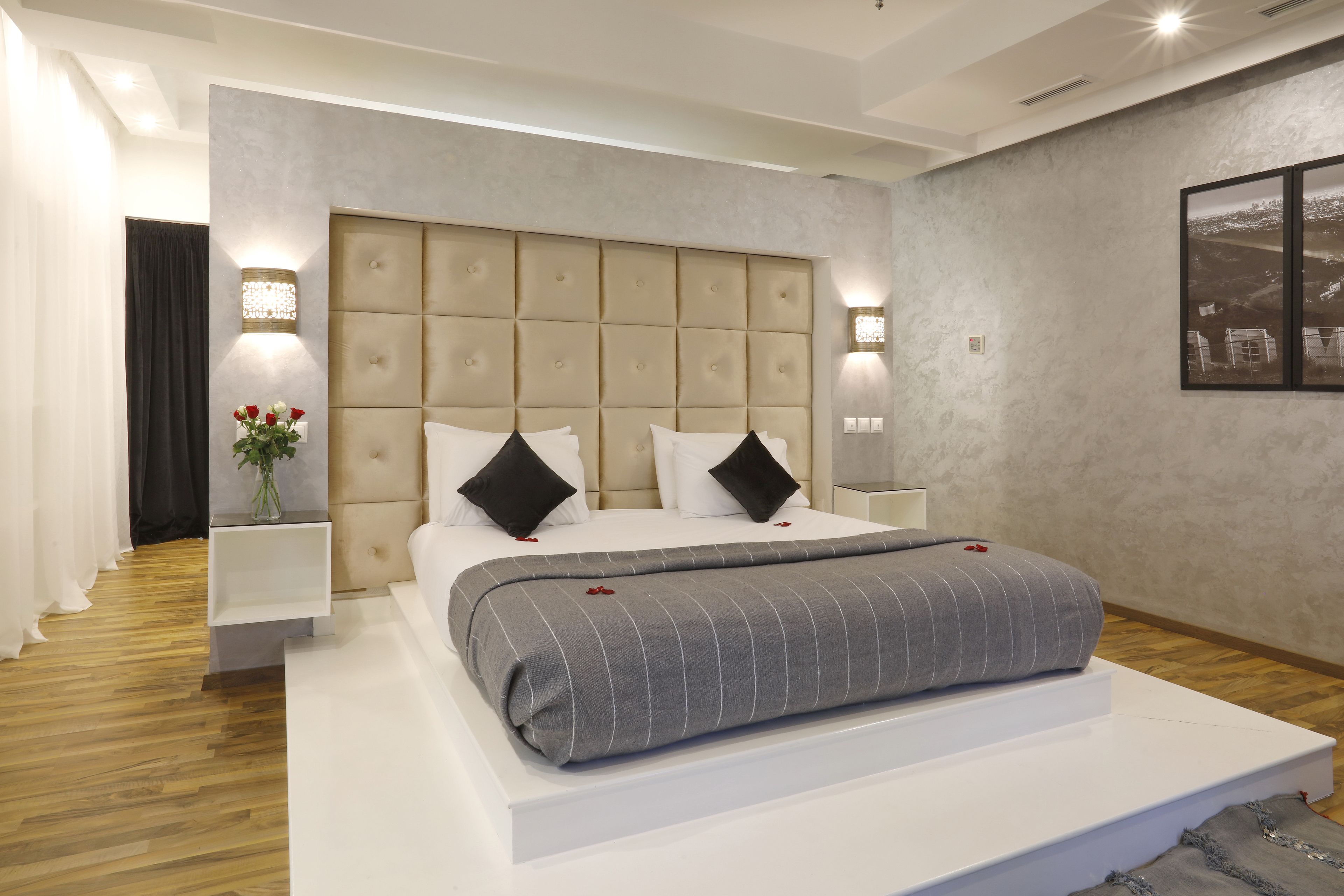 A modern bedroom with a tufted headboard, a large bed with a grey striped blanket, white pillows, and a vase of red flowers on a bedside table.