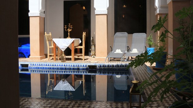 Zona piscina con pavimento piastrellato, due sdraio, un tavolo con tovaglia bianca e un candelabro decorativo.