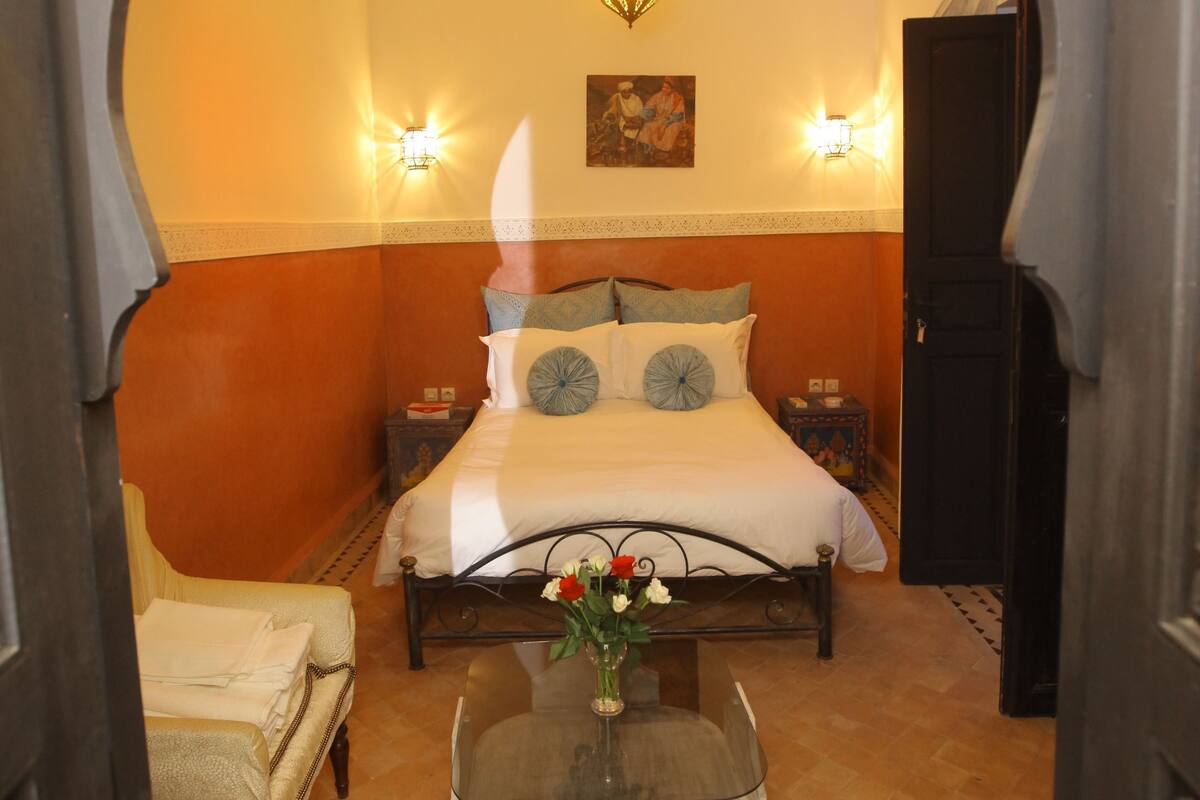A bedroom with a bed, a nightstand, a lamp, a chair, and a table with a vase.