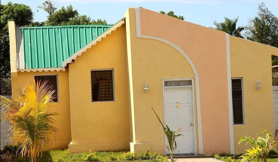 A single-story building with a yellow facade, a green roof, and a white door.