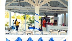 Un espace extérieur abrité, aménagé pour les repas, avec des tables et des chaises, un comptoir où sont présentés des plats, et une personne qui sert les clients.