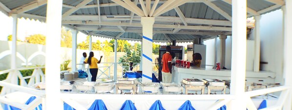 A covered outdoor dining area with tables and chairs, a counter with food items, and a person serving food.
