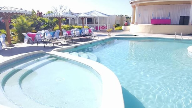 Une piscine entourée de chaises longues et de parasols, elle-même bordée de bâtiments aux couleurs blanches et roses.