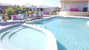 A swimming pool with lounge chairs and umbrellas, surrounded by buildings with white and pink accents.