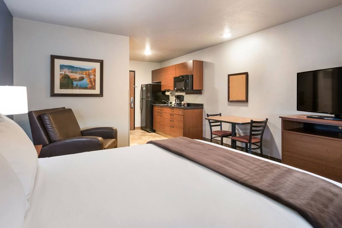 Ein Hotelzimmer mit einem Bett, einem Schreibtisch mit Stuhl, einem Fernseher, einer Küchenzeile und einem gerahmten Bild an der Wand.